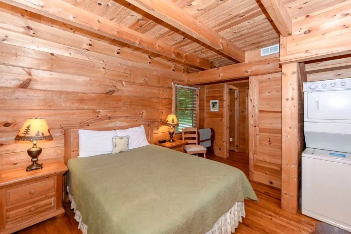 This image features a cozy wooden bed accented by soft lamps, with a washer and dryer set visible in the background. The room exudes warmth and comfort, with rustic wooden walls and a tranquil, restful ambiance. The presence of laundry appliances connects to guest convenience.