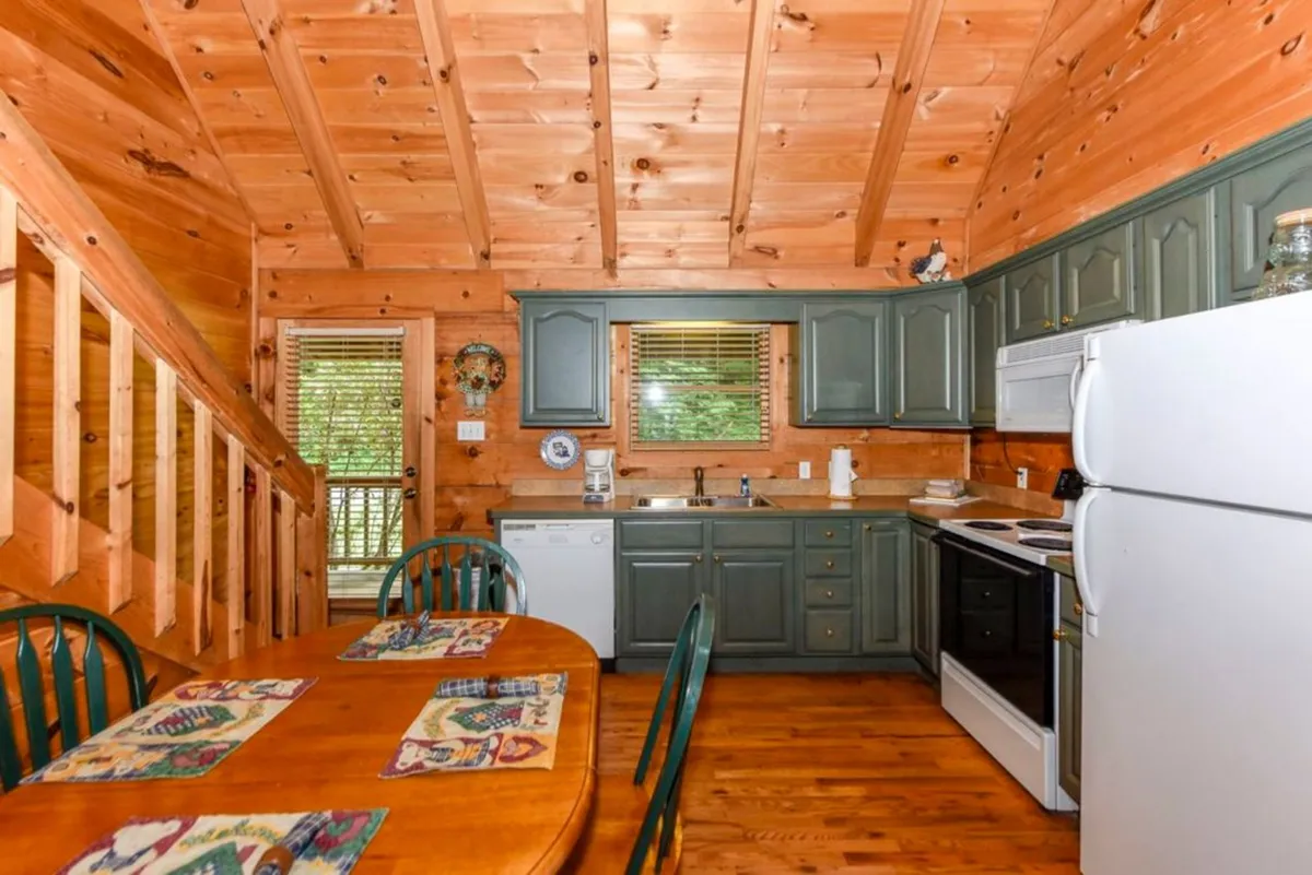 A spacious wooden dining table sits in front of green cabinets and a large window, with an open staircase in the background. The setting is bright and inviting, offering plenty of room for shared meals, board games, or simply enjoying the natural scenery outside. The kitchen is seamlessly connected to the dining area, highlighting the open-concept design.