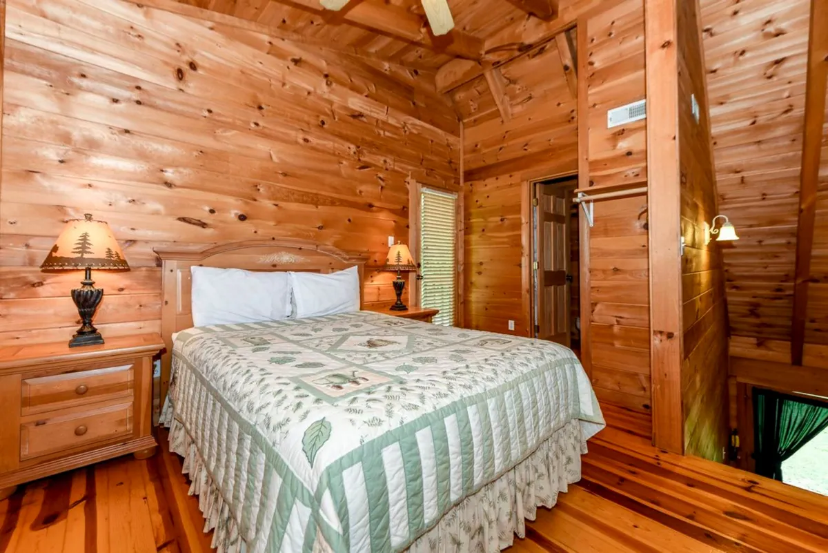 A sun-drenched bedroom with a wooden bed covered in a decorative quilt, a nightstand with a lamp, and authentic wood-paneled walls. The window allows natural light to filter in, highlighting the inviting and peaceful character of the space.