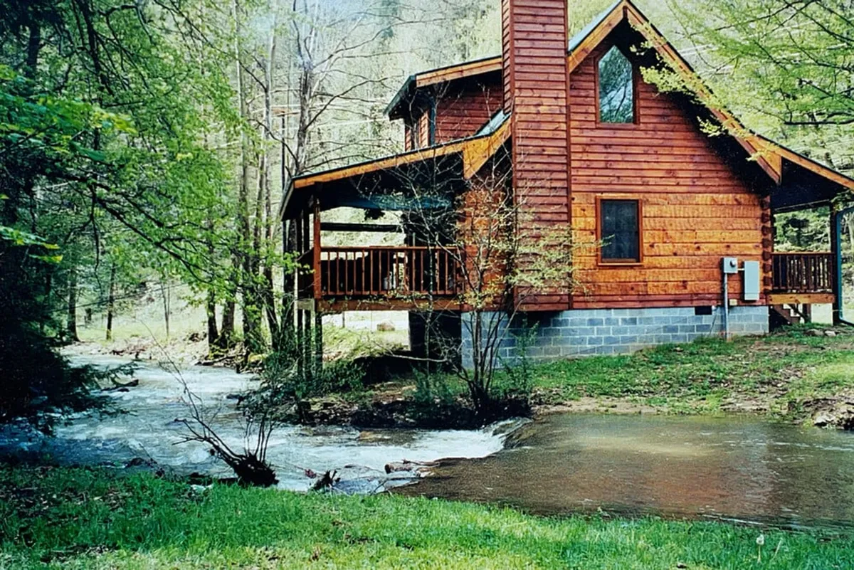This wide-angle shot displays the cabin’s deck overlooking a flowing creek, framed by tall trees and lush greenery. The image illustrates the seamless connection between the deck and the natural water feature, capturing moments of relaxation, birdwatching, and creekside enjoyment.