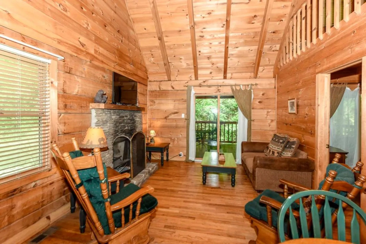 A welcoming great room with a stone fireplace as the centerpiece, flanked by large windows that let in woodland views. The space features wooden rocking chairs, a comfortable sofa, and a wooden coffee table, all arranged for easy conversation and relaxation. The warm firelight and natural textures create a cozy, inviting atmosphere ideal for gatherings.